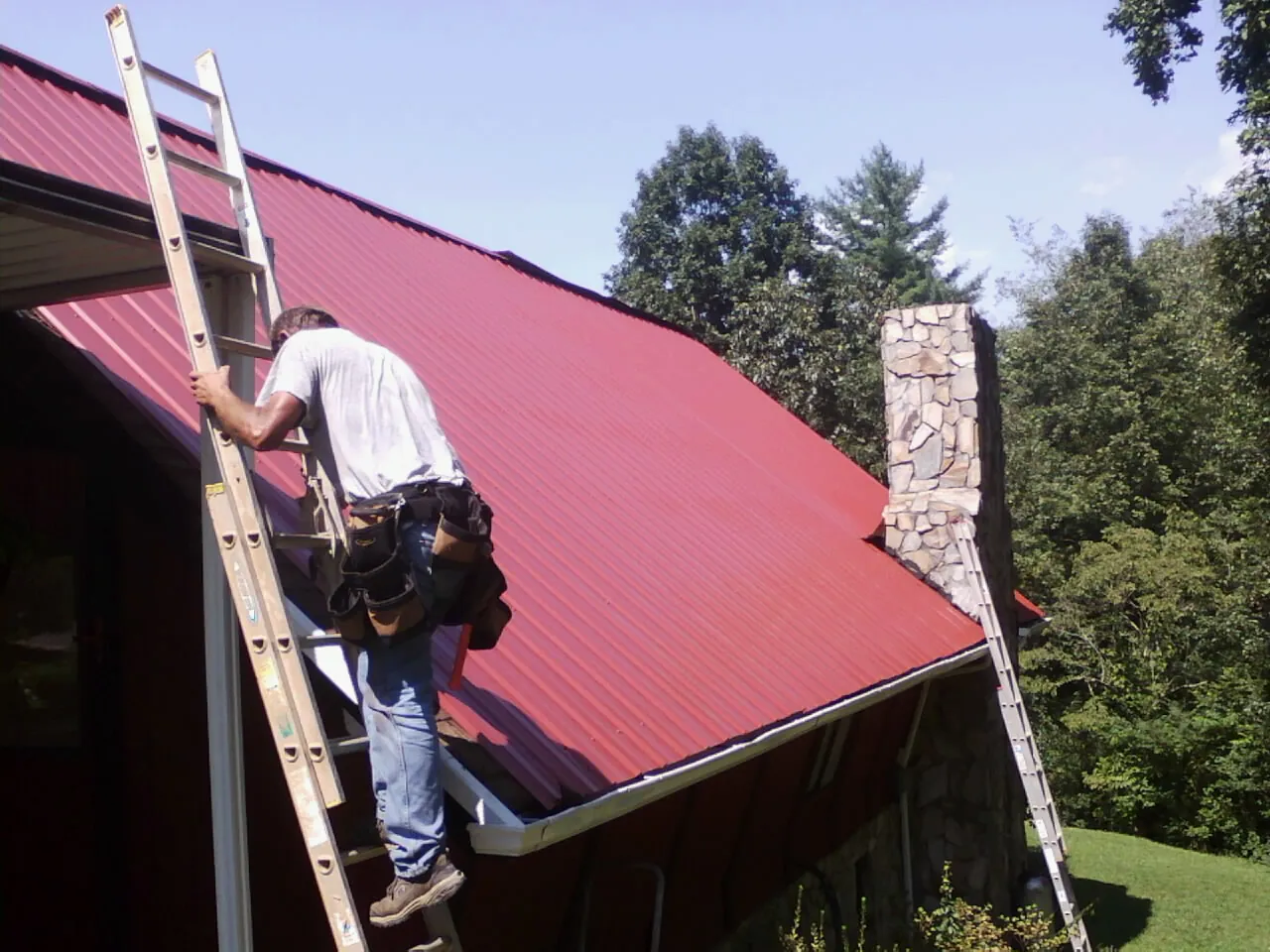 Roofer on a ladder beside a red metal roof and stone chimney — Soffit & Fascia Repair by Roofing Brothers in Pinehurst